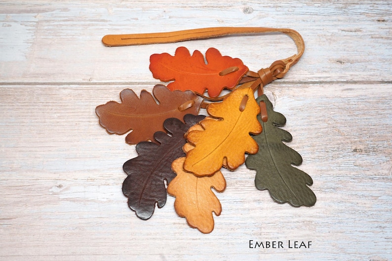 May include: A leather keychain featuring a cluster of six oak leaf-shaped cutouts in shades of orange, brown, yellow, and dark green. The leaves are attached to a thin leather strap with a loop. The text "EMBER LEAF" is printed in the corner.