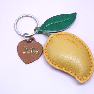 May include: A mango-shaped keychain with a green leaf and a brown heart-shaped tag. The mango is yellow with orange stitching. The heart tag has the name "Julie" engraved on it. The keychain is made of leather.