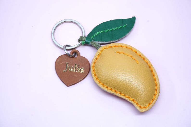 May include: A mango-shaped keychain with a green leaf and a brown heart-shaped tag. The mango is yellow with orange stitching. The heart tag has the name "Julie" engraved on it. The keychain is made of leather.