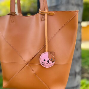 May include: A tan leather tote bag with a geometric design and stitched seams. The bag has two handles and a small, pink cat-shaped charm with brown accents hanging from one handle.