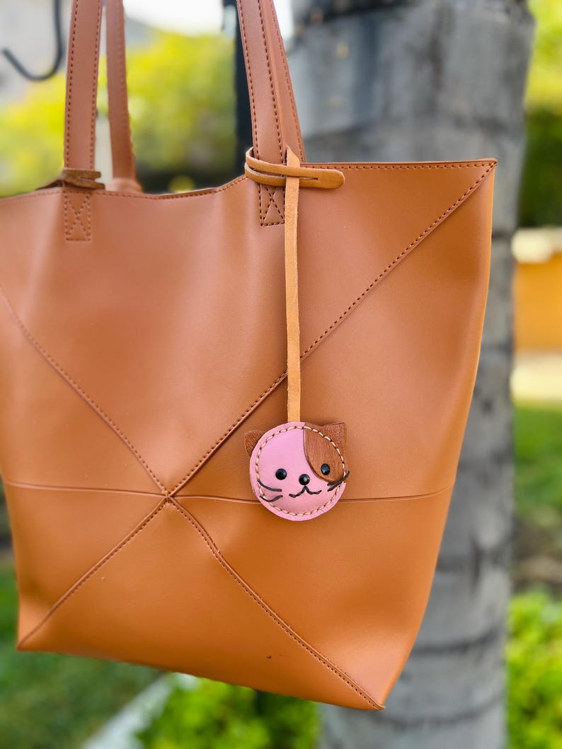 May include: A tan leather tote bag with a geometric design and stitched seams. The bag has two handles and a small, pink cat-shaped charm with brown accents hanging from one handle.