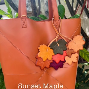 May include: A burnt orange tote bag featuring a smooth finish and stitched geometric paneling. A decorative cluster of leather maple leaf charms in shades of yellow, orange, red, purple, and green hangs from the handle. The text Sunset Maple is visible.
