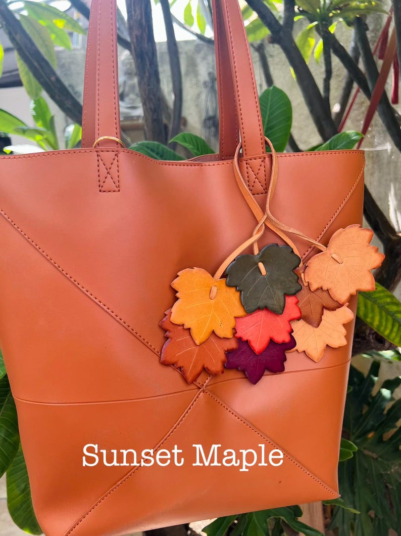 May include: A burnt orange tote bag featuring a smooth finish and stitched geometric paneling. A decorative cluster of leather maple leaf charms in shades of yellow, orange, red, purple, and green hangs from the handle. The text Sunset Maple is visible.