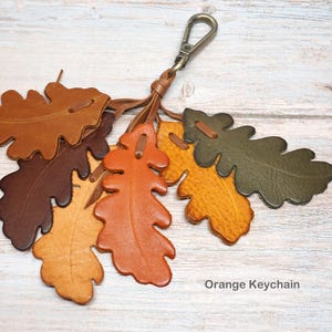 May include: A keychain featuring multiple oak leaf-shaped leather cutouts in shades of brown, orange, yellow, and olive green, attached to a metal lobster clasp. The text "Orange Keychain" is printed in the bottom right corner of the image.