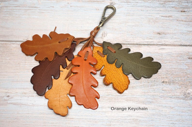 May include: A keychain featuring multiple oak leaf-shaped leather cutouts in shades of brown, orange, yellow, and olive green, attached to a metal lobster clasp. The text "Orange Keychain" is printed in the bottom right corner of the image.