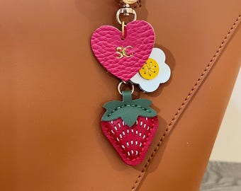 Puede incluir: Un llavero dorado con un dije de corazón de cuero rosa con las iniciales "SC" doradas, una flor blanca y amarilla, y un dije de fresa roja con hojas verdes.