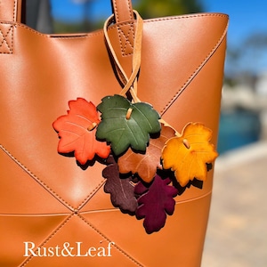 May include: A tan tote bag featuring a decorative charm made of multiple leather-like leaves in shades of orange, green, brown, yellow, and purple. The bag has stitched geometric paneling and the text Rust&Leaf printed in white on the front.