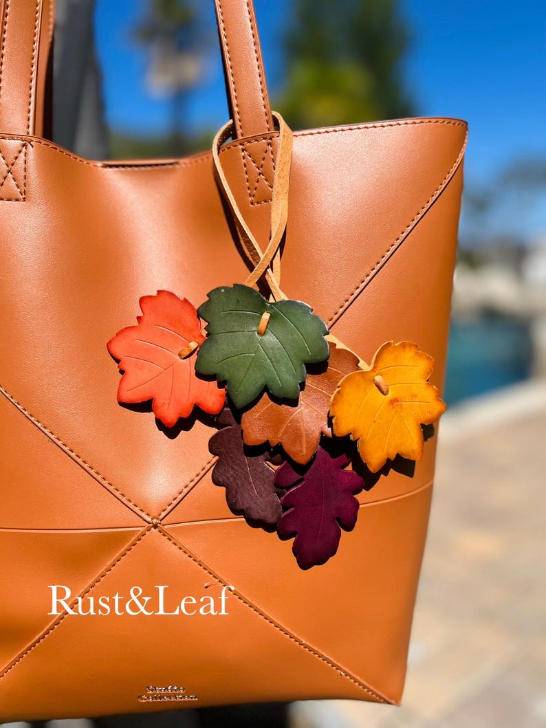 May include: A tan tote bag featuring a decorative charm made of multiple leather-like leaves in shades of orange, green, brown, yellow, and purple. The bag has stitched geometric paneling and the text Rust&Leaf printed in white on the front.