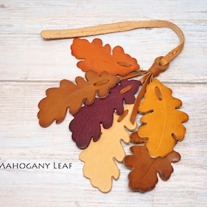 May include: A decorative hanging ornament featuring multiple oak leaf-shaped cutouts made of leather in shades of orange, yellow, deep red, and brown. The leaves are attached to a thin leather strap with a knot. The text MAHOGANY LEAF is visible.
