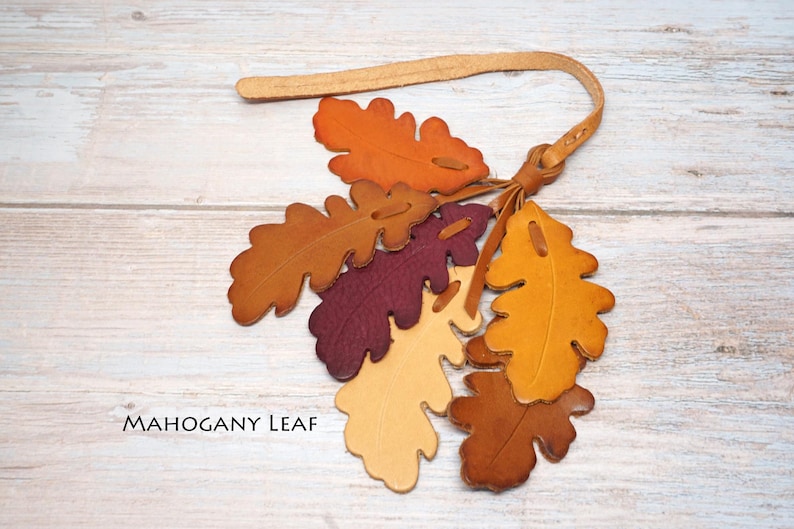 May include: A decorative hanging ornament featuring multiple oak leaf-shaped cutouts made of leather in shades of orange, yellow, deep red, and brown. The leaves are attached to a thin leather strap with a knot. The text MAHOGANY LEAF is visible.