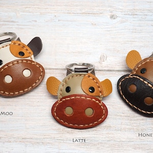 May include: Three cow-shaped keychains with metal rings. The left keychain, labeled Milly Moo, is white and brown. The center keychain, labeled Latte, is tan and reddish-brown. The right keychain, labeled Honey, is brown and black with tan ears.