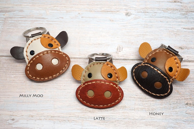May include: Three cow-shaped keychains with metal rings. The left keychain, labeled Milly Moo, is white and brown. The center keychain, labeled Latte, is tan and reddish-brown. The right keychain, labeled Honey, is brown and black with tan ears.