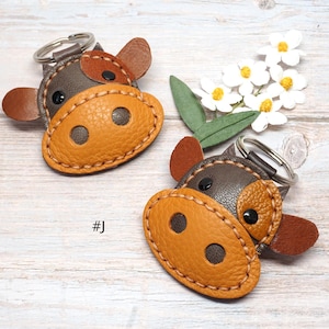 May include: Two cow-shaped keychains crafted from brown and tan textured leather with visible stitching. Each keychain features a dark brown head, a tan snout with two dark circular nostrils, two small ears, and black eyes, attached to a metal ring.
