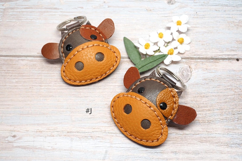 May include: Two cow-shaped keychains crafted from brown and tan textured leather with visible stitching. Each keychain features a dark brown head, a tan snout with two dark circular nostrils, two small ears, and black eyes, attached to a metal ring.