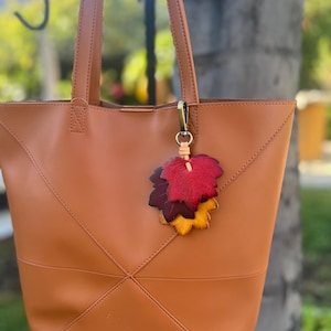 Könnte beinhalten: Eine hellbraune Ledertasche mit geometrischem Design, mit einem Cluster aus bunten Blattanhängern. Die Blätter sind in Rot-, Burgunder- und Gelbtönen gehalten und mit einem goldfarbenen Verschluss befestigt.