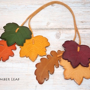 May include: A decorative garland featuring multiple leather leaves in shades of green, yellow, orange, brown, and deep red, all strung together on a thin tan leather cord. The text AMBER LEAF is printed in black in the bottom left corner.