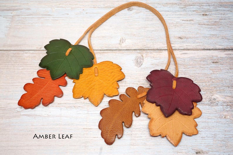 May include: A decorative garland featuring multiple leather leaves in shades of green, yellow, orange, brown, and deep red, all strung together on a thin tan leather cord. The text AMBER LEAF is printed in black in the bottom left corner.