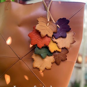 May include: A tan leather tote bag features a hanging charm made of multiple leather leaves in shades of orange, yellow, green, brown, and purple. The text "Imperfect charm" is printed in white at the bottom of the bag.