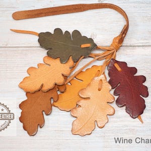 May include: A leather strap featuring six oak leaf-shaped cutouts in shades of olive green, tan, light brown, and deep burgundy. Each leaf is attached to the strap with a thin leather cord. A logo reads GENUINE LEATHER BY BASHA LEATHER.