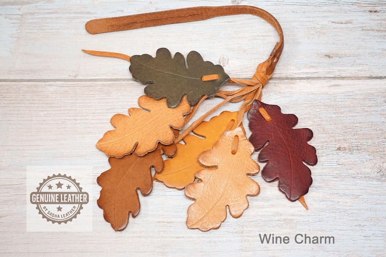 May include: A leather strap featuring six oak leaf-shaped cutouts in shades of olive green, tan, light brown, and deep burgundy. Each leaf is attached to the strap with a thin leather cord. A logo reads GENUINE LEATHER BY BASHA LEATHER.
