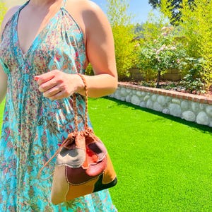 May include: A patchwork leather drawstring bag with a braided strap. The bag features various colors, including brown, tan, red, and black. The bag is held by a person wearing a turquoise floral dress.