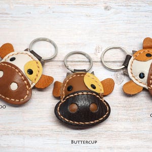 May include: Three cow-shaped keychains made of leather with metal rings. The left keychain is labeled TOFFEE MOO, featuring a white and brown face. The center keychain is labeled BUTTERCUP, with a dark brown face. The right keychain is labeled COOKIE.