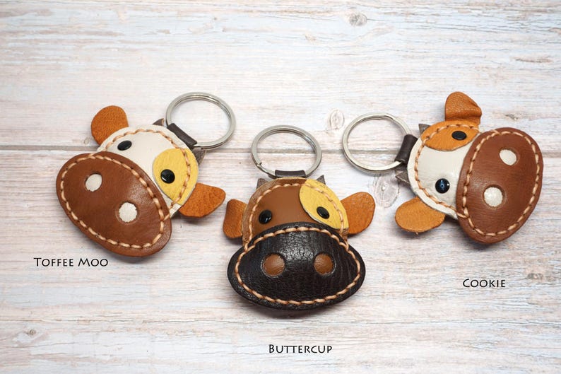 May include: Three cow-shaped keychains made of leather with metal rings. The left keychain is labeled TOFFEE MOO, featuring a white and brown face. The center keychain is labeled BUTTERCUP, with a dark brown face. The right keychain is labeled COOKIE.