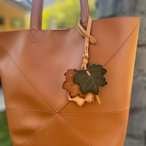 May include: A tan tote bag with a leaf-shaped charm. The bag has a geometric design with stitched seams and a Studio Collection logo. The charm features three leather leaves in shades of brown and green, attached to a leather strap.