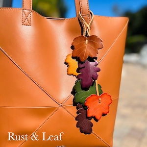 May include: A tan tote bag with a leather leaf charm featuring multiple colors including brown, yellow, purple, green, and orange. The bag has stitched geometric lines and the text Rust & Leaf printed in white on the lower front section.