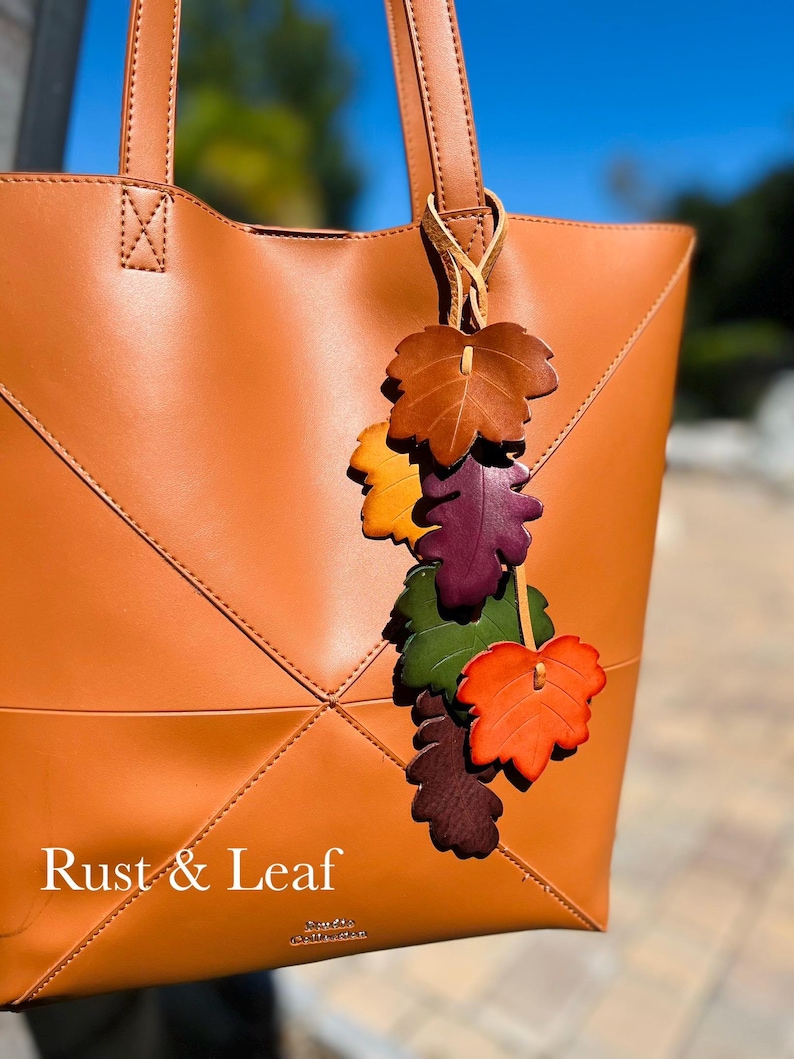 May include: A tan tote bag with a leather leaf charm featuring multiple colors including brown, yellow, purple, green, and orange. The bag has stitched geometric lines and the text Rust & Leaf printed in white on the lower front section.