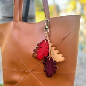 May include: A tan leather tote bag with two shoulder straps and a decorative charm featuring three leather leaves in red, tan, and dark burgundy. The charm is attached to the bag with a bronze-colored metal lobster clasp. The word Red is visible.
