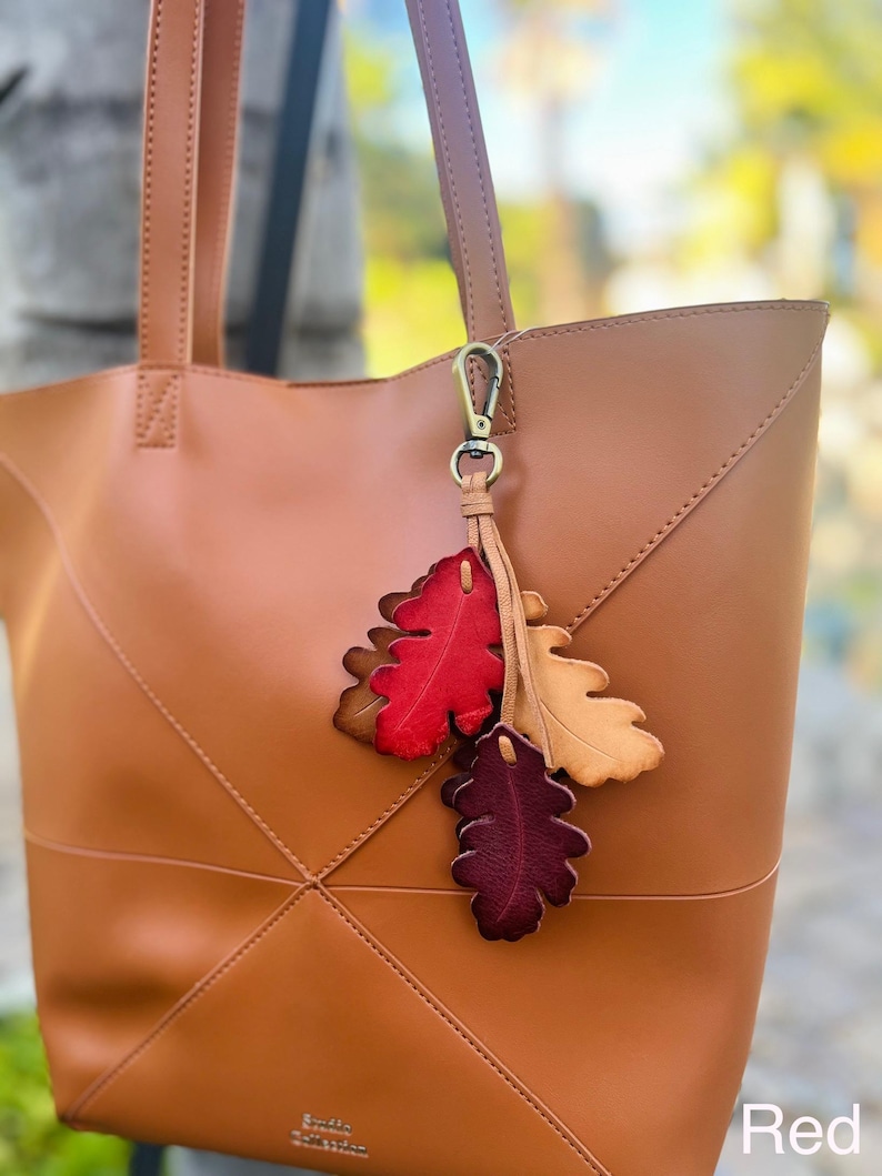 May include: A tan leather tote bag with two shoulder straps and a decorative charm featuring three leather leaves in red, tan, and dark burgundy. The charm is attached to the bag with a bronze-colored metal lobster clasp. The word Red is visible.