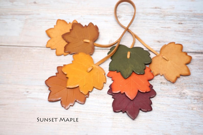 May include: A collection of leather maple leaf cutouts in shades of yellow, orange, brown, green, and deep red, attached to a thin leather cord. The text SUNSET MAPLE is printed in black in the lower left corner of the image.