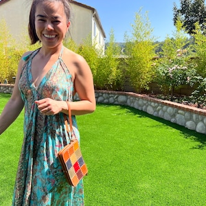 Peut inclure: Une femme porte une longue robe turquoise à imprimé floral et tient un petit sac à main en cuir patchwork avec une sangle marron. En arrière-plan, une pelouse verte, un mur en pierre et une maison sous un ciel bleu clair.