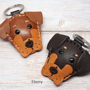May include: Two leather dog-shaped keychains with metal rings. One keychain is tan with a lighter tan muzzle, and the other is ebony with a tan muzzle. Both feature stitched details, black circular eyes, and a black nose. Text labels read Tan and Ebony.