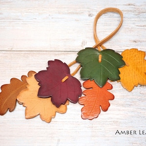 May include: A decorative hanging ornament featuring six leather leaves in shades of brown, yellow, burgundy, green, and orange, attached to a thin leather loop. The text AMBER LEAF is printed in the bottom right corner of the image.