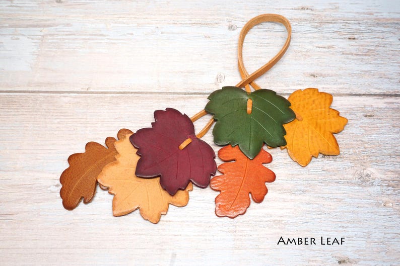May include: A decorative hanging ornament featuring six leather leaves in shades of brown, yellow, burgundy, green, and orange, attached to a thin leather loop. The text AMBER LEAF is printed in the bottom right corner of the image.