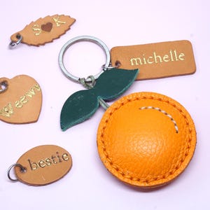 May include: A collection of leather keychains in various shapes and colors. Includes a heart, oval, leaf, and rectangular tag, all with gold lettering. One keychain is orange and round with white stitching. The keychains are on a light purple background.