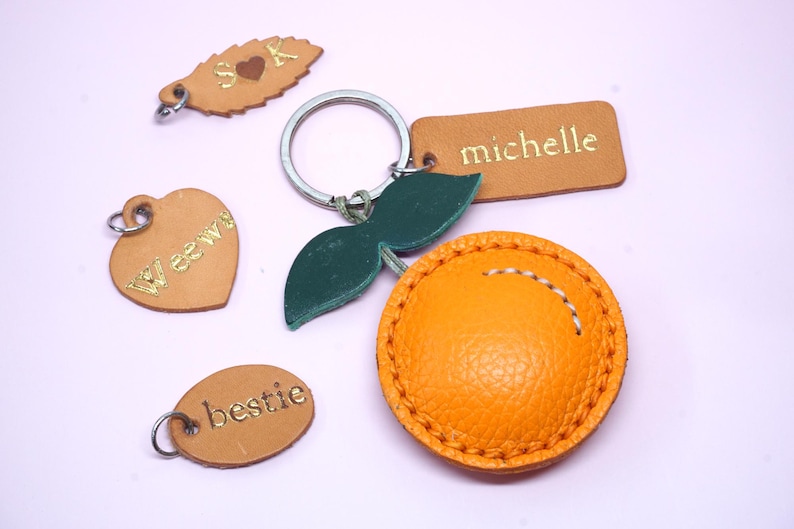 May include: A collection of leather keychains in various shapes and colors. Includes a heart, oval, leaf, and rectangular tag, all with gold lettering. One keychain is orange and round with white stitching. The keychains are on a light purple background.