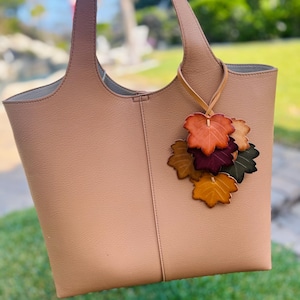 May include: A light tan tote bag with two shoulder straps and a vertical seam down the center. Attached to one strap is a decorative charm featuring a cluster of multi-colored leather leaves in shades of orange, yellow, burgundy, and dark green.