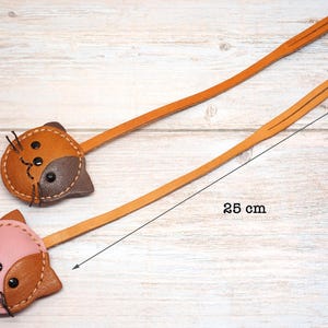May include: Two leather cat-shaped bookmarks. One is brown and tan, the other is pink and tan. Each bookmark has a long, thin, tan leather strap. The length of the bookmark is 25 cm.