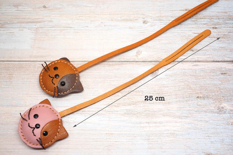 May include: Two leather cat-shaped bookmarks. One is brown and tan, the other is pink and tan. Each bookmark has a long, thin, tan leather strap. The length of the bookmark is 25 cm.