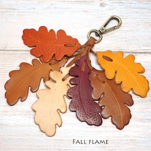 May include: A leather keychain featuring a cluster of six oak leaf-shaped cutouts in shades of orange, brown, cream, burgundy, and yellow. The leaves are attached to a bronze-toned metal lobster clasp. The text FALL FLAME is printed below.