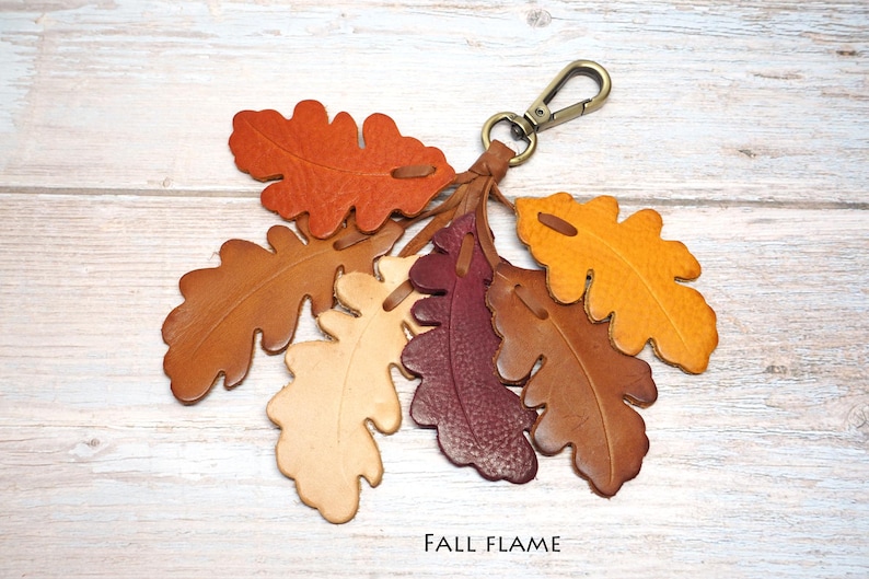 May include: A leather keychain featuring a cluster of six oak leaf-shaped cutouts in shades of orange, brown, cream, burgundy, and yellow. The leaves are attached to a bronze-toned metal lobster clasp. The text FALL FLAME is printed below.