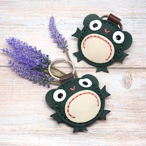 May include: Two dark green leather keychains shaped like frogs with cream-colored bellies, white and black eyes, and red stitched details. Each keychain features a metal ring attached by a brown leather tab, showcasing visible dark green stitching.
