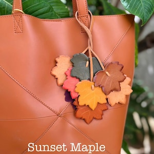 May include: A tan tote bag featuring a decorative cluster of leather leaf charms in shades of green, red, orange, and brown. The bag has stitched geometric paneling and the text "Sunset Maple" printed in white at the bottom center.