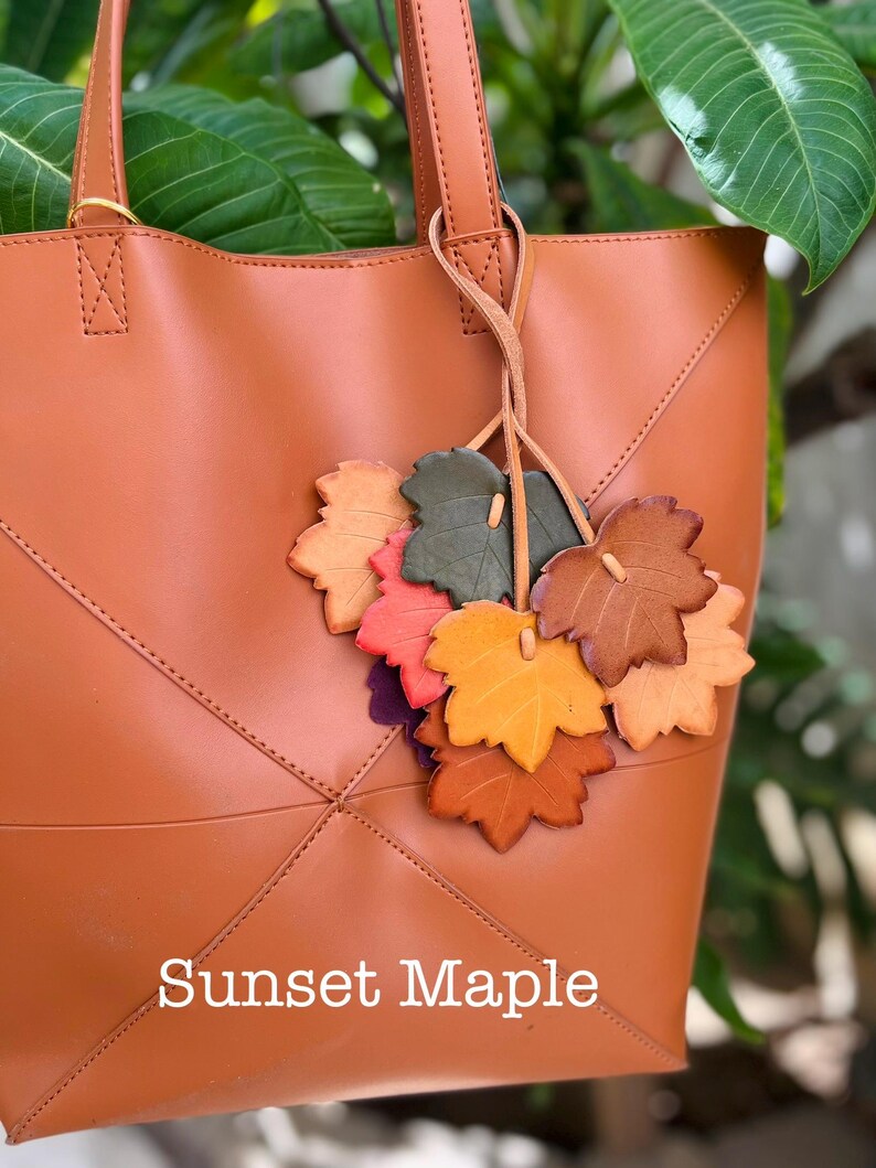 May include: A tan tote bag featuring a decorative cluster of leather leaf charms in shades of green, red, orange, and brown. The bag has stitched geometric paneling and the text "Sunset Maple" printed in white at the bottom center.
