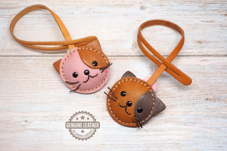 May include: Two leather cat-shaped keychains. One is pink and brown, the other is brown and dark brown. Both have black eyes and stitched details. Each keychain has a loop for attaching to keys. The words "GENUINE LEATHER" are printed on the image.