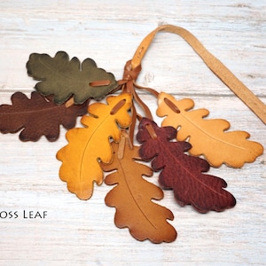 May include: A collection of six leather oak leaf cutouts in shades of olive green, dark brown, golden yellow, tan, and deep burgundy, all attached to a single leather strap. The text MOSS LEAF is printed in black in the lower left corner.