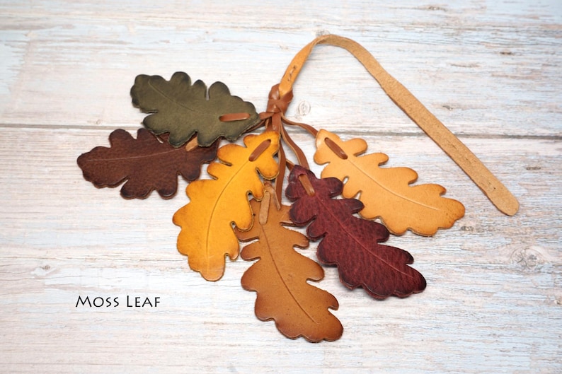 May include: A collection of six leather oak leaf cutouts in shades of olive green, dark brown, golden yellow, tan, and deep burgundy, all attached to a single leather strap. The text MOSS LEAF is printed in black in the lower left corner.